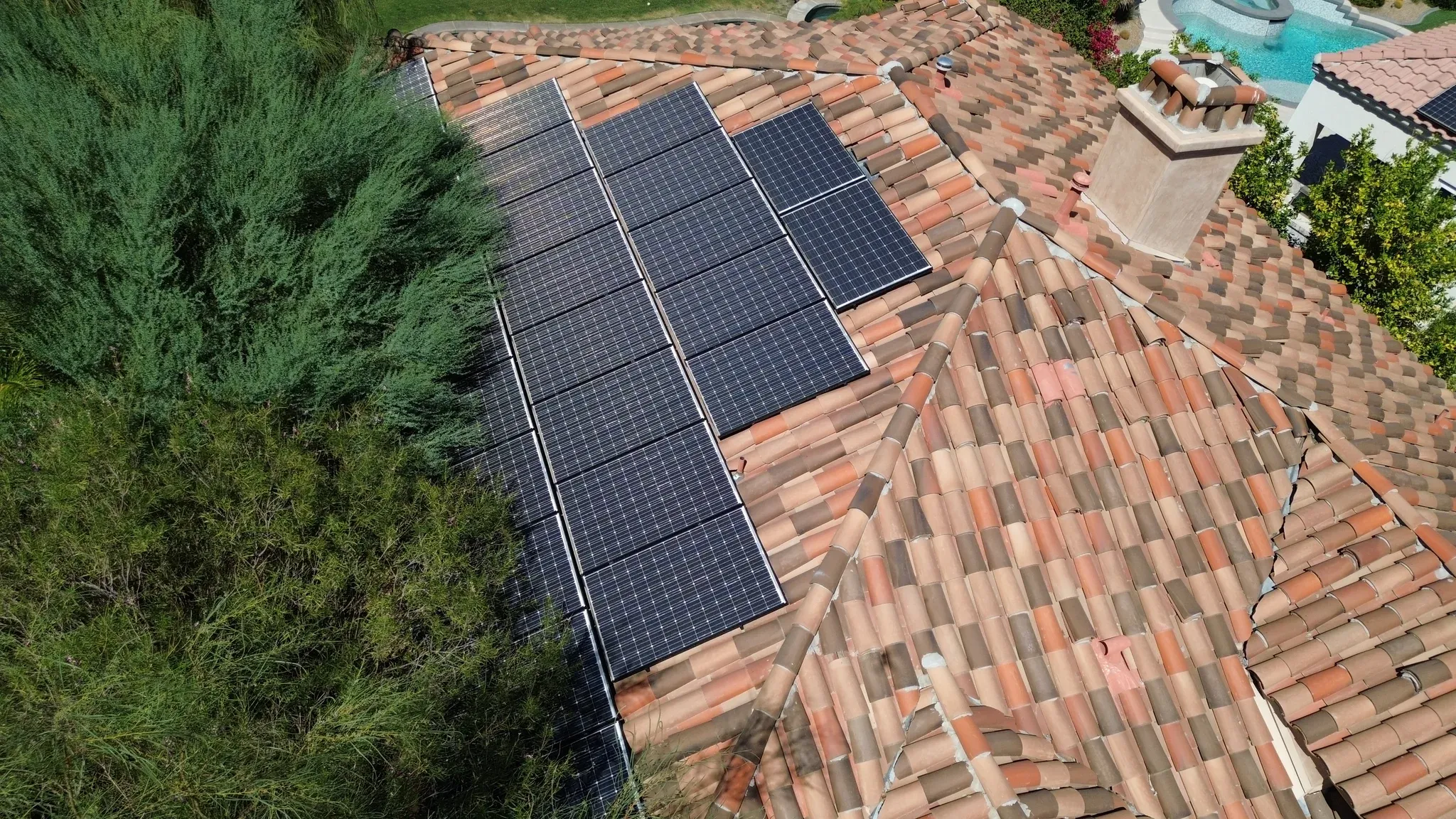 Drone view of a long solar panel run on a multicolor clay tile roof edged by desert shrubs, with a pool in the background