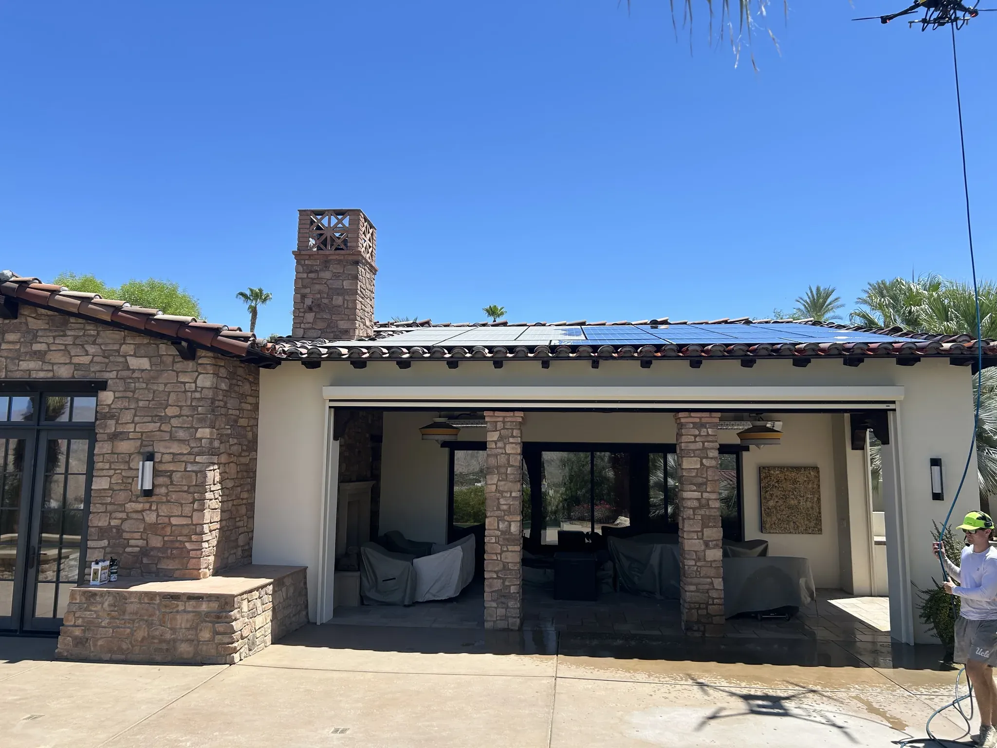 DronePower1 cleaning drone hovering above a Spanish tile roof in the Coachella Valley while a technician on the patio below feeds the water line during a solar panel cleaning