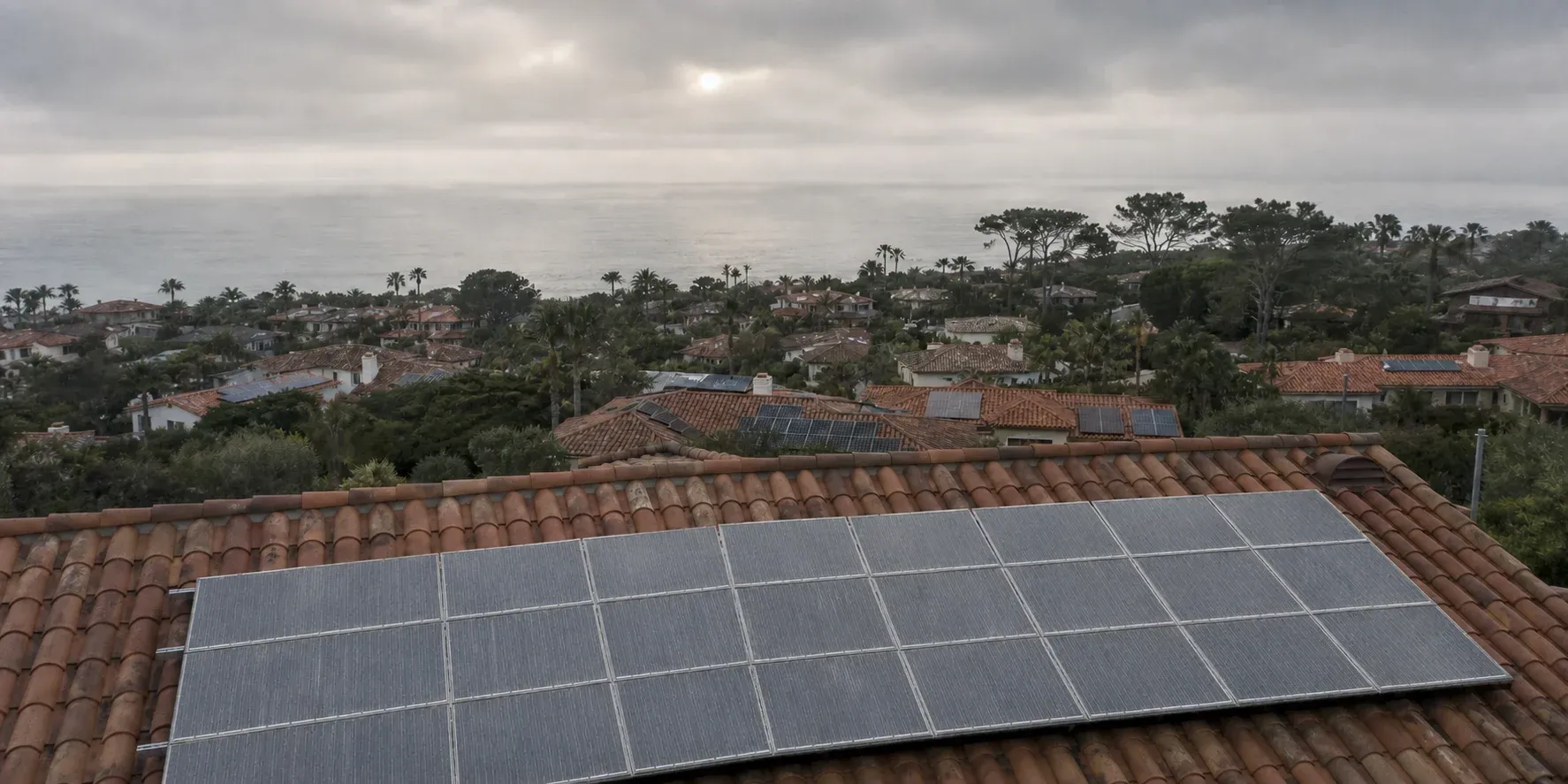 Marine layer over a coastal North County neighborhood of tile roofs at dawn, foreground rooftop solar panels dulled by a pale salt haze, the Pacific flat and grey behind