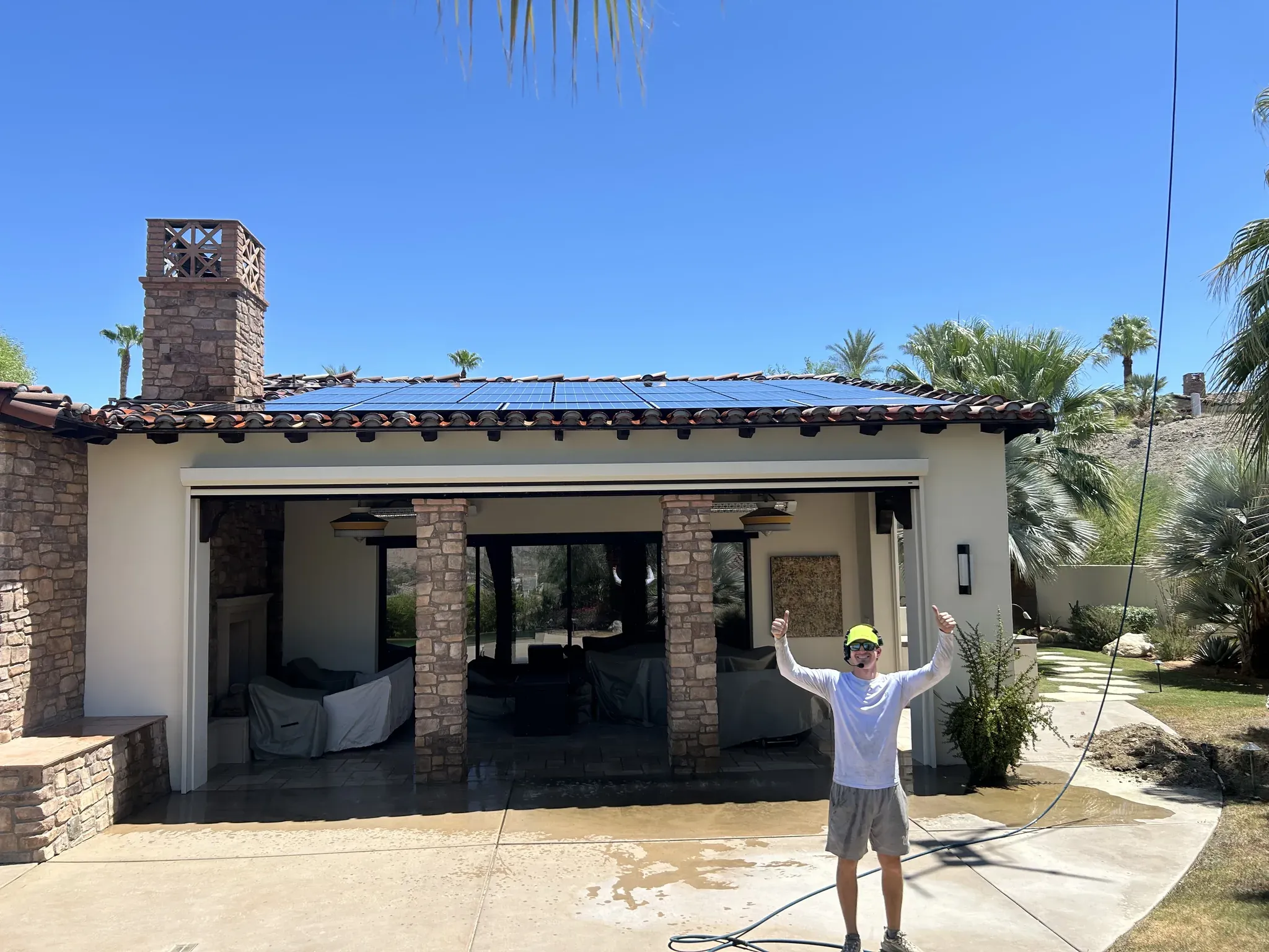 DronePower1 solar cleaning technician in a hi-vis cap and headset raising both arms in front of a freshly cleaned rooftop solar array on a Coachella Valley tile-roof home