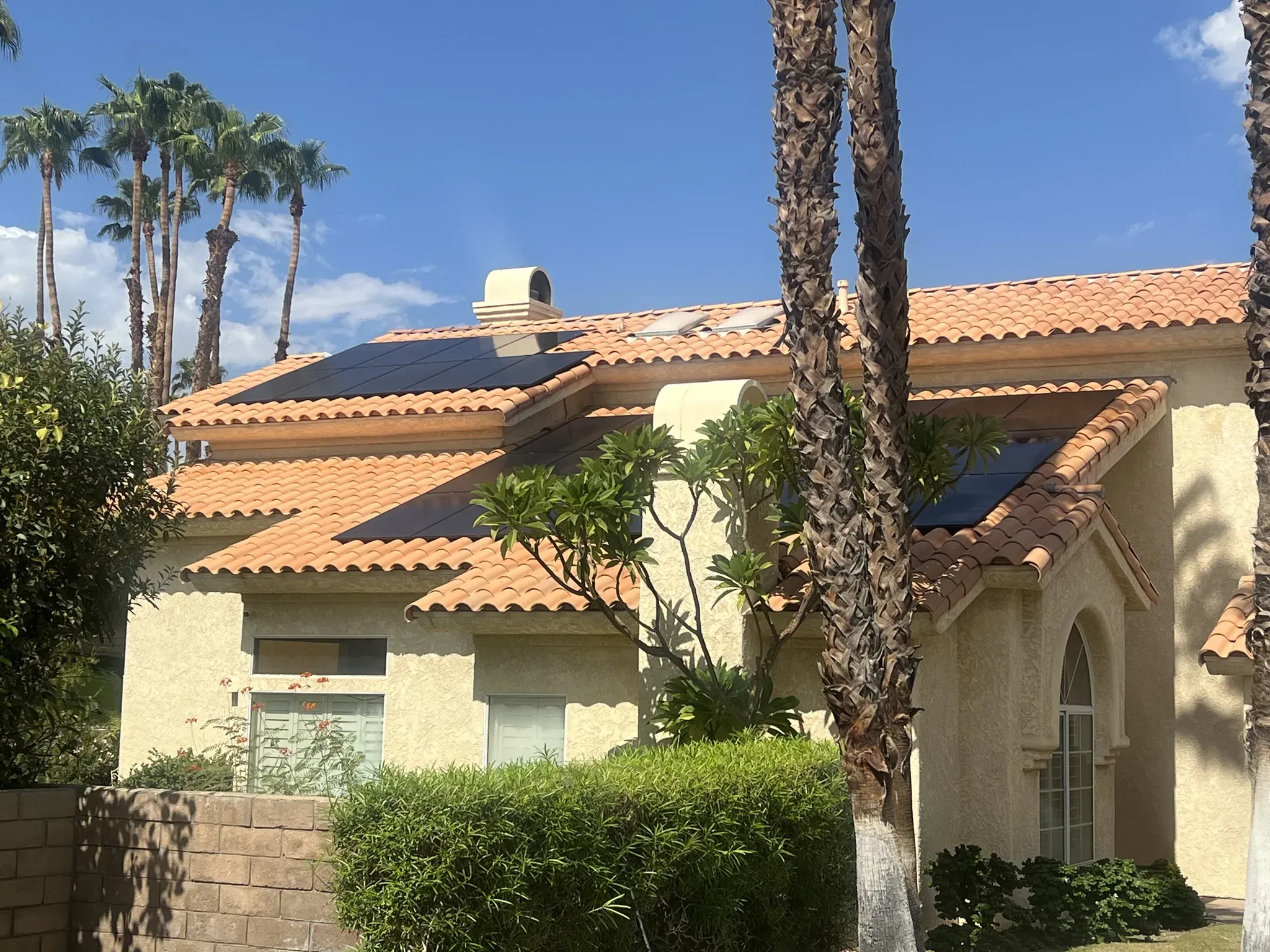 Street-level view of a stucco home with dark solar panels across its terracotta tile roof, framed by palm trees under a blue sky